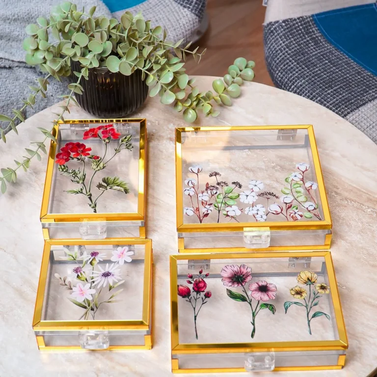 Set of four acrylic floral keepsake boxes with gold edges displayed on a round marble table with a plant decor.