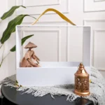 Rectangular acrylic gift carrier with mustard leather handle holding decorative figurine and lantern.