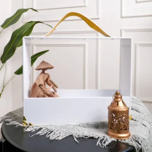 Rectangular acrylic gift carrier with mustard leather handle holding decorative figurine and lantern.